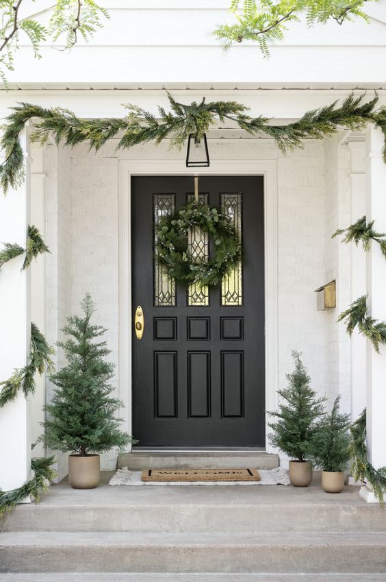a farmhouse Christmas porch with an evergreen garland and wreath, potted Christmas trees and layered rugs