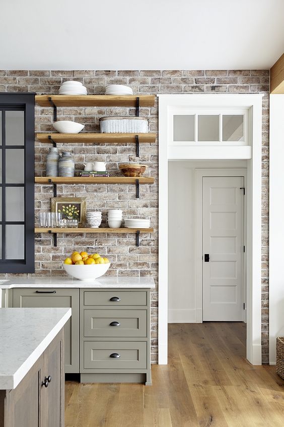 a farmhouse kitchen with a brick wall, olive green cabinets, open shelving and a transom window located above the door