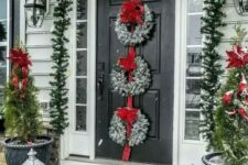 a festive Christmas porch with an evergreen garland with lights and red ribbons, snowy wreaths with red bows and mini Christmas trees