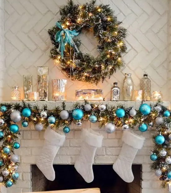 a fir garland with blue and silver ornaments plus lights, candles in mercury glass candleholders and a greenery wreath with lights and a turquoise bow