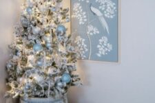 a flocked Christmas tree in a planter, with pearly, silver and pastel blue ornaments, a pastel blue bow on top and some lights