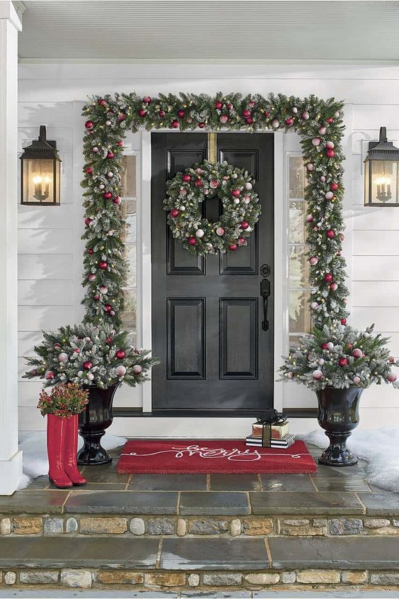 a glam Christmas porch styled with an evergreen garland with white and red ornaments, a matching wreath and arrangements in pots plus red rubber boots
