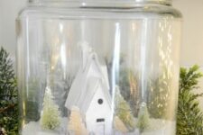 a large jar Christmas terrarium with faux snow, mini trees and a white house surrounded with lights and evergreens