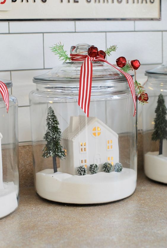 a large jar with faux snow, a mini tree and a lit up house topped with red bells and greenery is cool for Christmas
