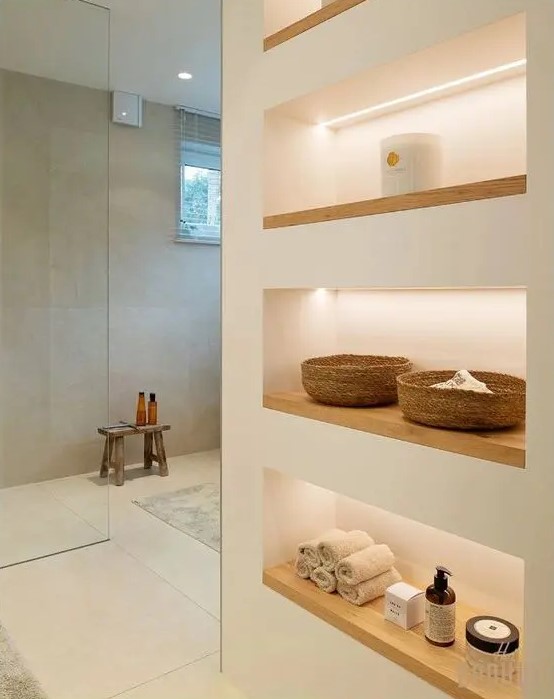 a minimalist bathroom with neutral walls, a large shower space enclosed in glass, a series of lit up niche shelves for storage