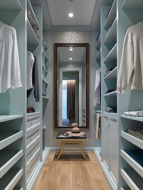 a narrow blue closet with open storage compartments, drawers and railings, a mirror and a black leather stool