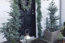 a natural Christmas porch with flocked Christmas trees in baskets, candle lanterns, mini trees and an evergreen garland over the door