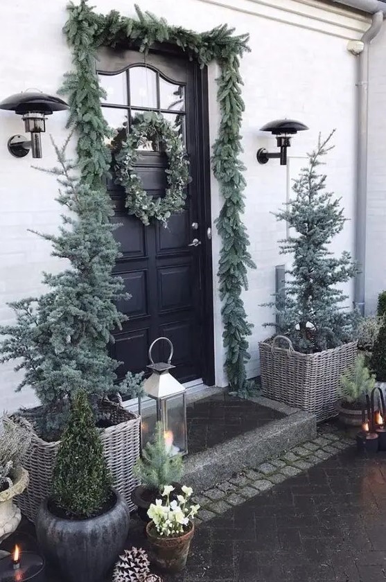 a natural Christmas porch with flocked Christmas trees in baskets, candle lanterns, mini trees and an evergreen garland over the door