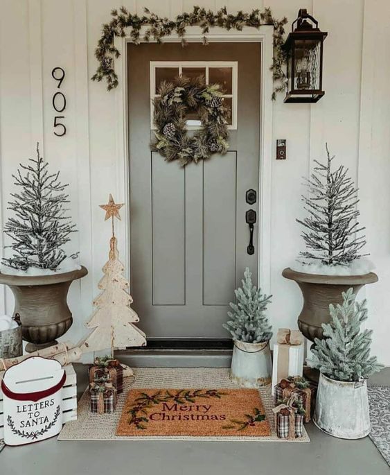 a pretty Christmas porch with potted Christmas trees, wooden trees, firewood, an evergreen garland and wreath, gift boxes