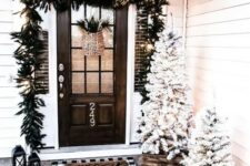 a pretty farmhouse Christmas porch with snowy Christmas trees in crates, an evergreen garland with lights over the door, candle lanterns
