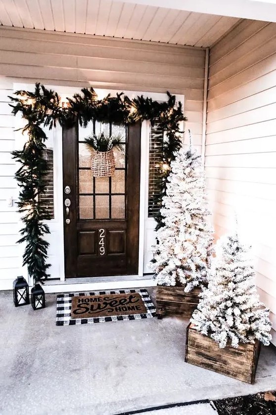 a pretty farmhouse Christmas porch with snowy Christmas trees in crates, an evergreen garland with lights over the door, candle lanterns