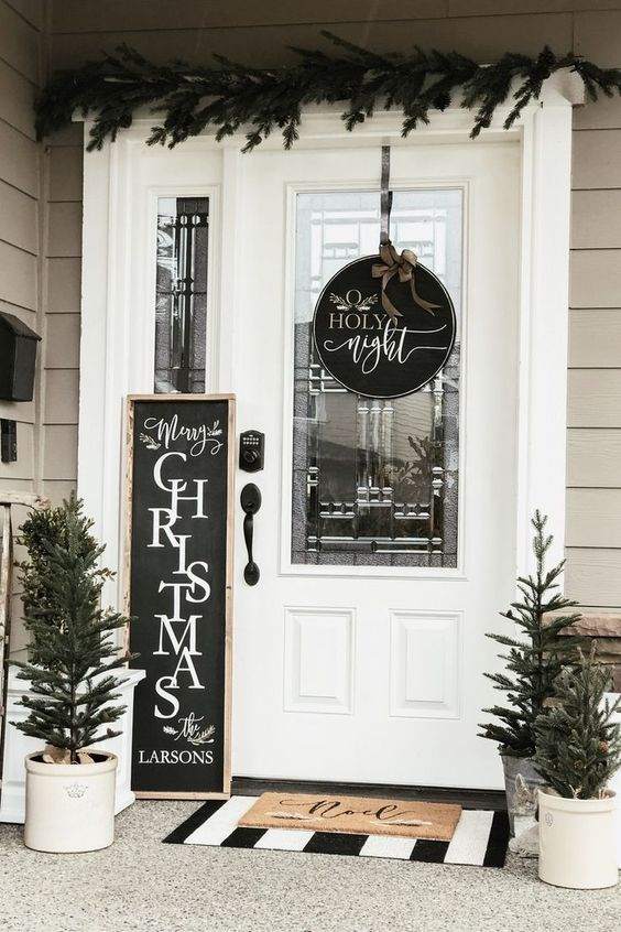a rustic black and white Christmas porch with a couple of signs, potted Christmas trees and an evergreen garland over the door