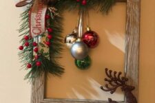 a rustic frame Christmas wreath with evergreens, berries, a burlap bow and some colorful ornaments plus a sparkling deer