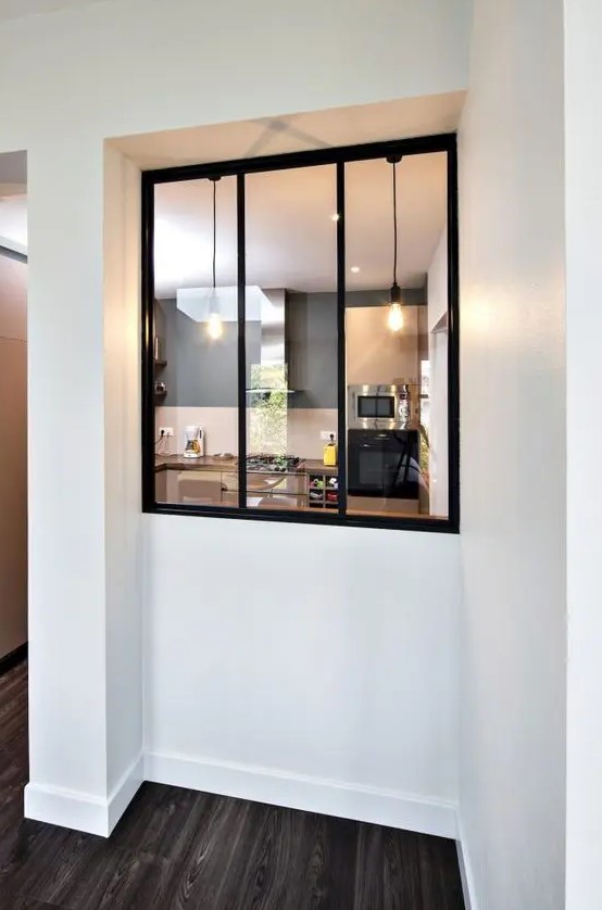 a simple interior window with black framing connects the kitchen and the entryway and lets enjoy the warmth and coziness of the kitchen