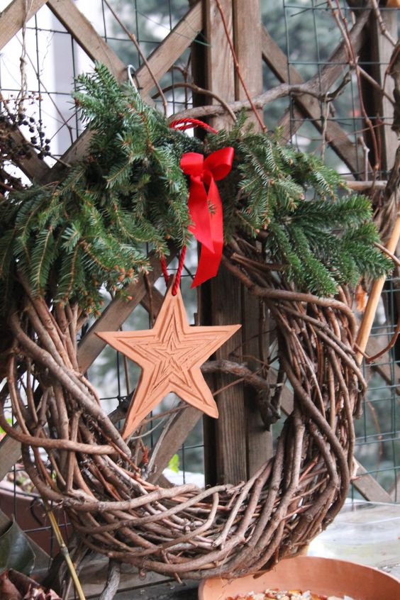 a simple rustic Christmas wreath of grapevine, evergreens, a wooden star and a red bow is a stylish and cozy idea