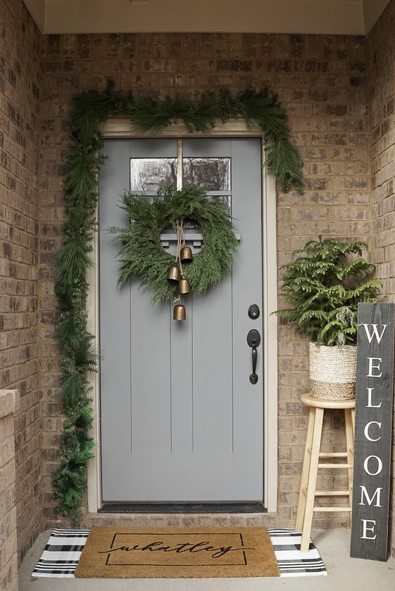a small and cozy Christmas porch with an evergreen garland and wreath, a potted plant, a black sign and vintage bells