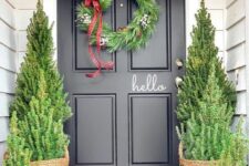 a stylish Christmas porch done with Christmas trees in baskets and a matching wreath with a plaid ribbon bow is a cool idea