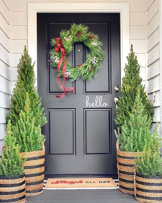 a stylish Christmas porch done with Christmas trees in baskets and a matching wreath with a plaid ribbon bow is a cool idea