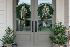 a stylish farmhouse Christmas porch with evergreen wreaths with bells, potted Christmas trees, candle lanterns