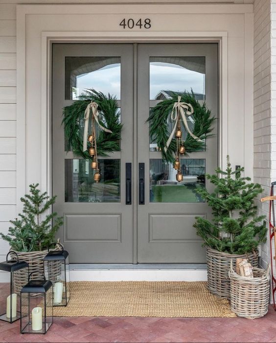 a stylish farmhouse Christmas porch with evergreen wreaths with bells, potted Christmas trees, candle lanterns