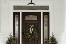 a stylish modern farmhouse Christmas porch with potted Christmas trees, an evergreen garland and a wreath, firewood and wooden candle lanterns