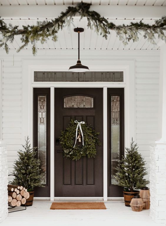 a stylish modern farmhouse Christmas porch with potted Christmas trees, an evergreen garland and a wreath, firewood and wooden candle lanterns