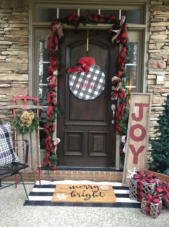 a vintage farmhouse Christmas porch with plaid ribbons, evergreens, a plaid sign, vine gifts and pillows plus a decorated sleigh