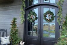 a welcoming and cozy Christmas porch with an evergreen garland and wreaths and potted Christmas trees, firewood in buckets