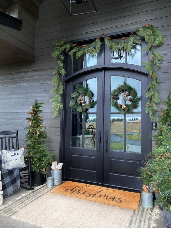 a welcoming and cozy Christmas porch with an evergreen garland and wreaths and potted Christmas trees, firewood in buckets