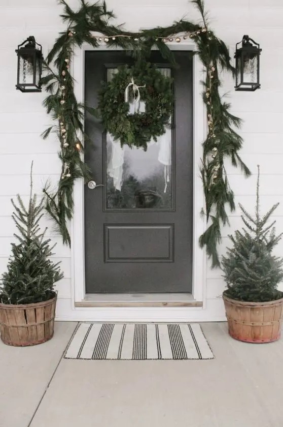 a woodland Christmas porch with mini trees in wooden buckets, a fir garland with metallic ornaments and a fir wreath on the foor is very cool