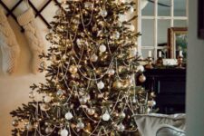an elegant and gold-fashioned Christmas tree decorated with white and metallic ornaments, beads and lights plus leaves on top