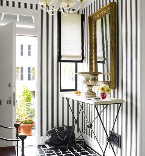 an elegant and chic entryway with a black and white floor, black and white stripe wallpaper on the walls, black and white blinds and a vintage frame mirror