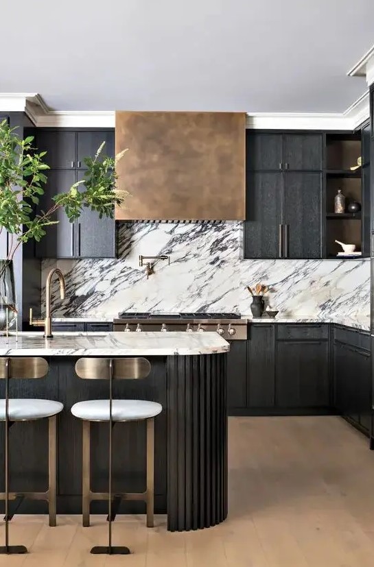 a sophisticated black kitchen with a white marble backsplash, an aged brass hood, a fluted curved kitchen island and tall stools with brass backs