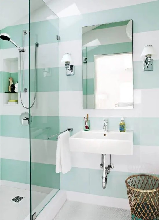 a contemporary bathroom with mint and white striped walls, a small shower space and a wall mounted sink, a mirror and a basket