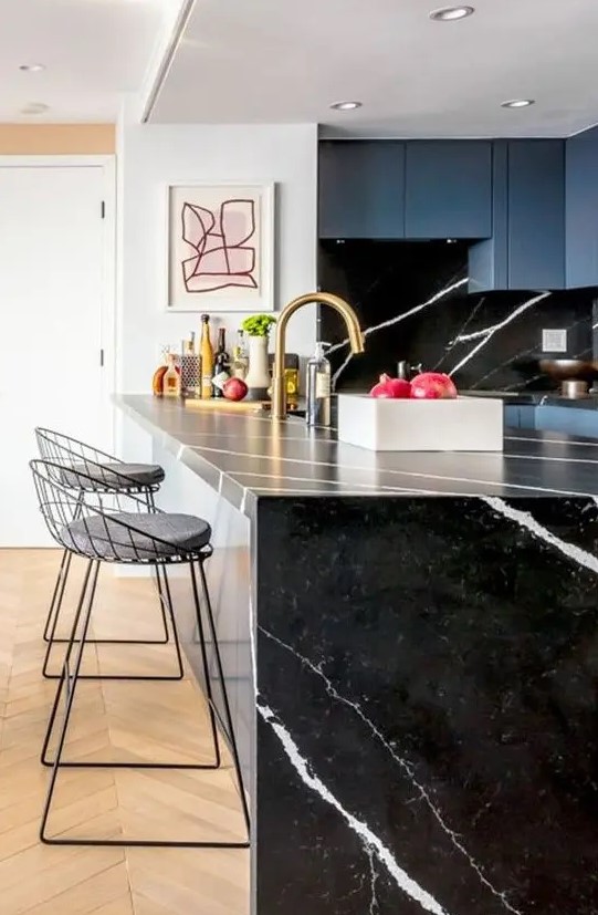 a blue kitchen with a black marble backsplash and a waterfall black countertop plus touches of gold for a chic look