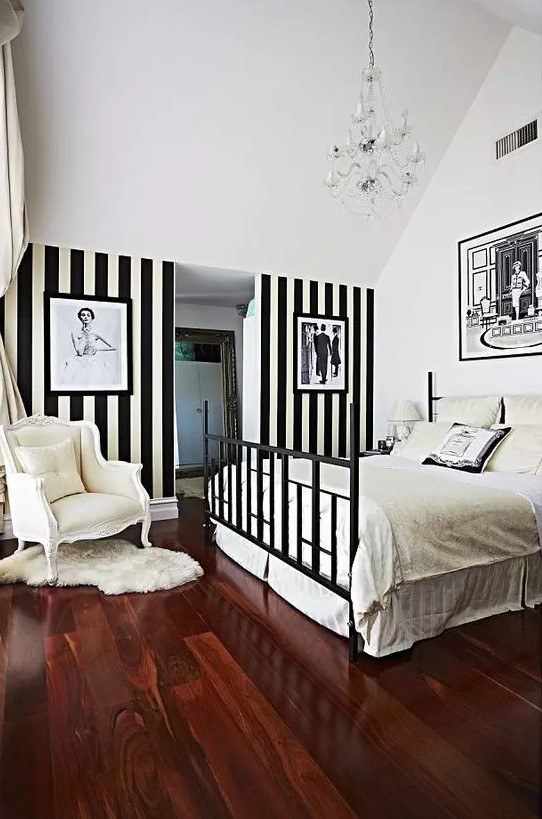 a refined vintage bedroom with a black bed and neutral bedding, an accent black and white stripe wall, a neutral chair and a crystal chandelier
