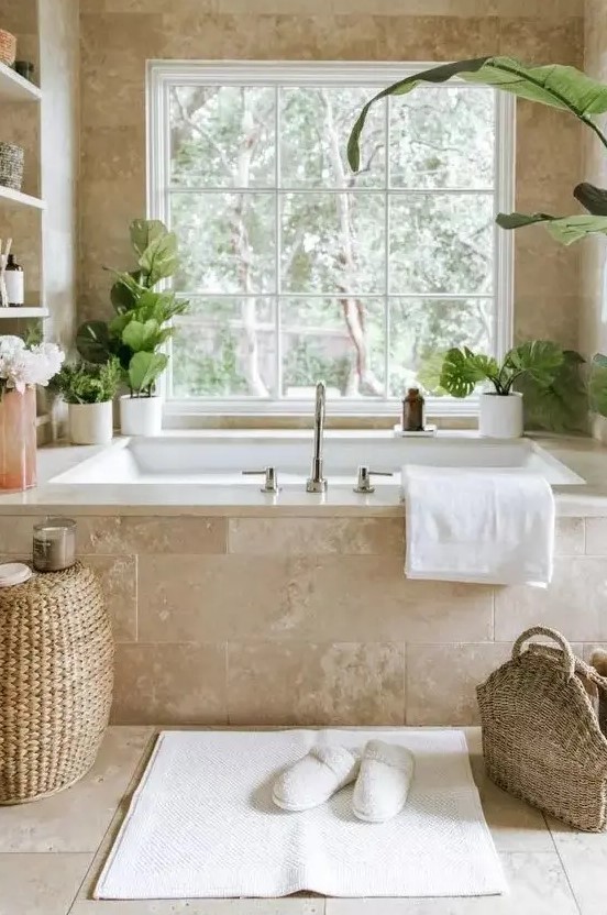 a gorgeous neutral spa bathroom clad with limestone tiles, with a tub clad with them, built in shelves and potted greenery, a woven pouf and a bag