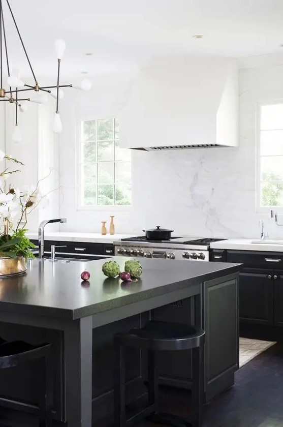 a cool black kitchen with a large white hood, a large black kitchen island and matching stools, a chandelier and some brass touches