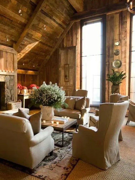 a gorgeous chalet living room with rough wooden walls and a ceiling, a fireplace clad with stone, neutral furniture, greenery and blooms