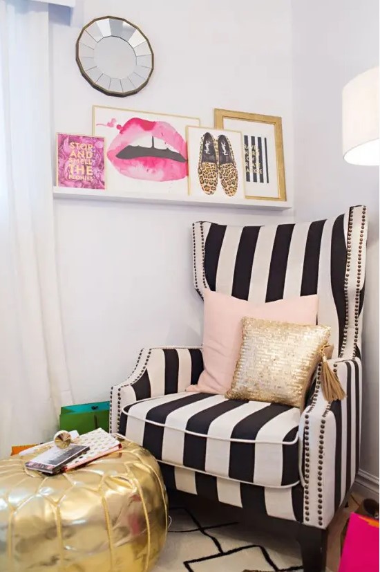 a glam corner with a black and white stripe wingback chair, some pink and gold pillows, a ledge gallery wall and a gold pouf