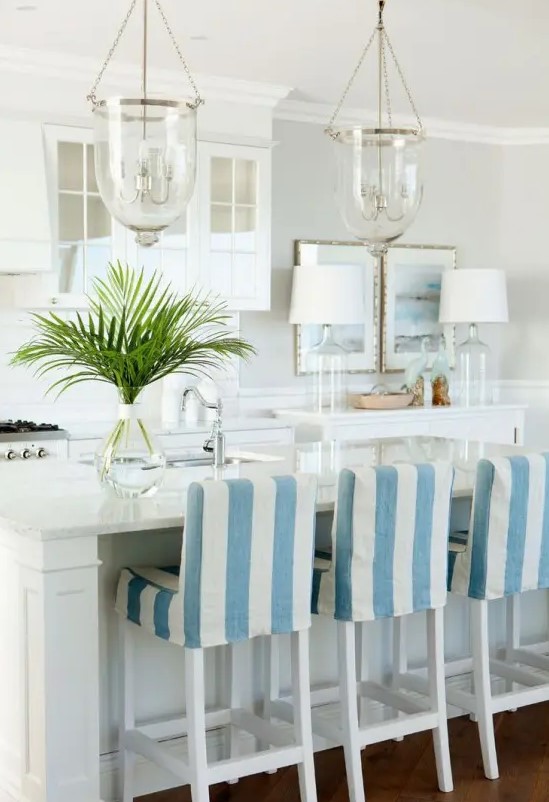 a neutral coastal kitchen with white cabinets, glass and usual ones, a console table with decor, a kitchen island and tall blue and white stools