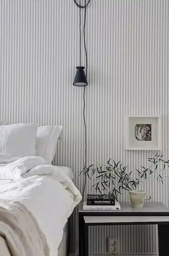 a Scandinavian bedroom finished off with a grey and white striped wall, a bed and a nightstand, some greenery and art is welcoming