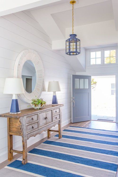 a seaside entryway with a shabby chic wooden console table, blue lamps and a round mirror in a whitewashed frame, a striped rug