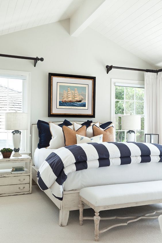 a beach bedroom with a whitewashed bed with striped bedding, shabby chic nightstands and a lovely bench plus a ship artwork