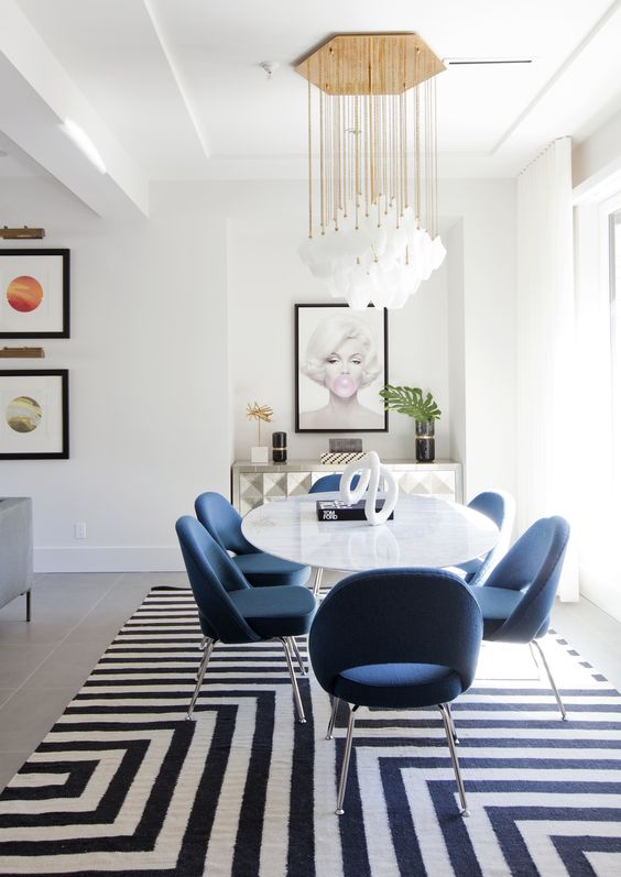 a gorgeous dining space with a quirky credenza, an oval table and blue chairs, a statement geometric rug and a cool chandelier