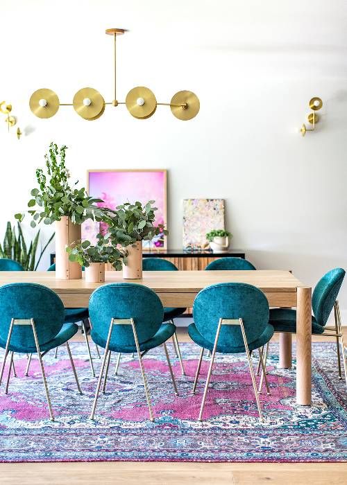 a colorful dining room with a dining table in neutral stain, teal chairs and a pink artwork plus a rug that echoes with both chairs and art