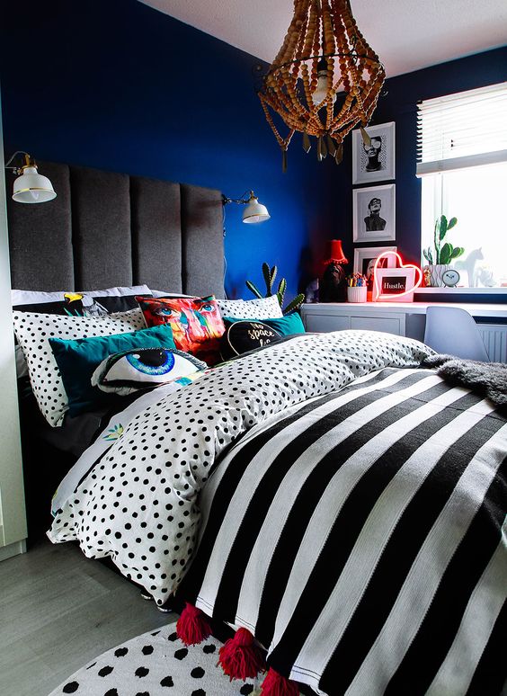 a gorgeous moody bedroom with blue walls, an upholstered bed with polka dot and striped bedding, a beaded chandelier