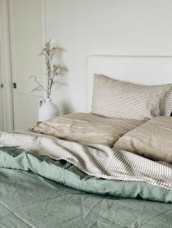 a neutral bedroom with a neutral bed, neutral and striped bedding, a green duvet, some grasses in a vase