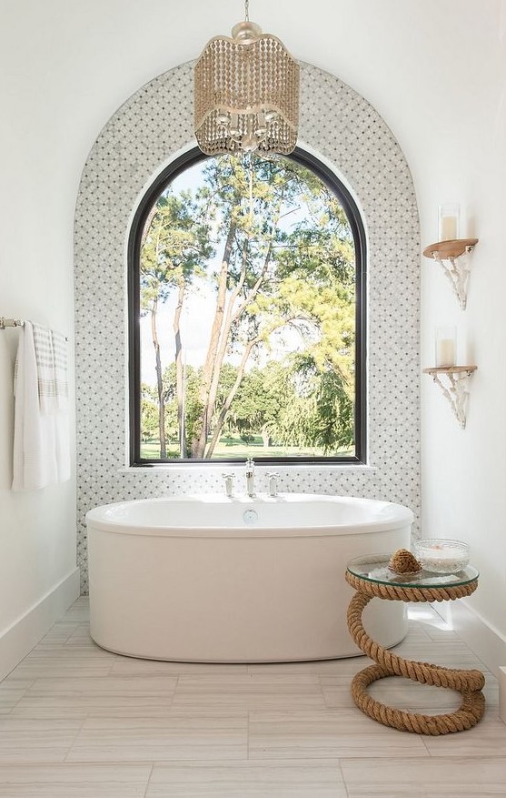 a bold bathroom with marble tiles and an arched window for an accent, an oval tub, a beaded chandelier and a rope side table