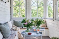 a Scandinavian sunroom clad with white beadboard, with a built-in bench and pastel textiles, blue tables and blooms